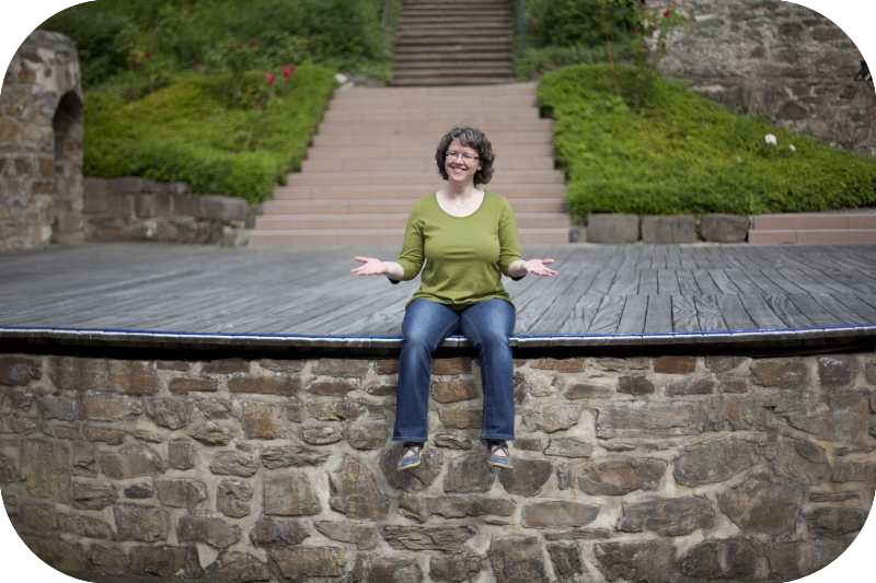 Daniela Wilhelm sitzt am Rand einer Bühne und hält die Handflächen offen nach oben und lächelt.